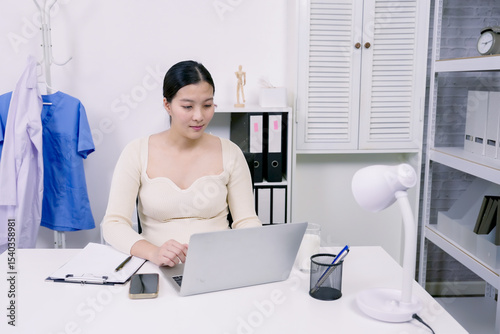 Asian pregnant woman sitting at office desk typing on laptop surrounded by work tools showing professionalism productivity and balance between career and health in final stage of pregnancy