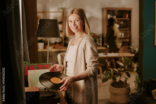 Caucasian senior woman smiling while holding vinyl record near turntable, standing in cozy living room with another senior in background, engaging in relaxed home activity