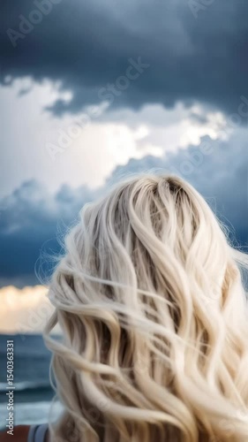 Woman with long platinum blonde wavy hair gazing at ocean under dramatic cloudy sky, creating serene and contemplative mood.