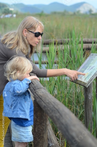 Grandmother educates her granddaughter about swamp plants, sharing knowledge and pointing to an information board. The scene emphasizes learning and bonding in nature