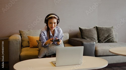 An adult asian woman enjoys her leisure time immersed in gaming by holding the joystick control via laptop on a comfortable sofa at cozy living room atmosphere. Her headphones enhance the fun