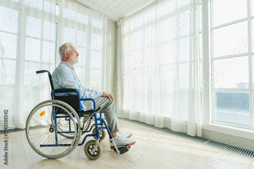 Sad senior man sitting in wheelchair looking out window in retirement home hospital. Old paralyzed man in chair for people with disability feel depressed. Lonely elderly sick man sitting on wheelchair