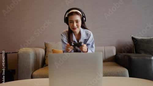 An adult asian woman enjoys her leisure time immersed in gaming by holding the joystick control via laptop on a comfortable sofa at cozy living room atmosphere. Her headphones enhance the fun