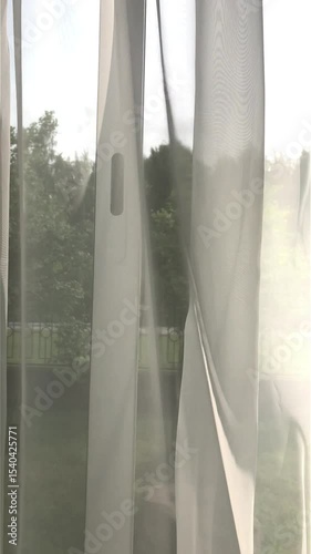 Curtains on the window are blown by the wind. The view from the room to the outside through the windows.