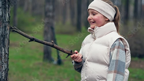 Happy Caucasian teenage girl plays in autumn forest holding and waving stick she striking tree trunk. She is smiling and laughing during her funny game. Adventurous pastime in the park concept
