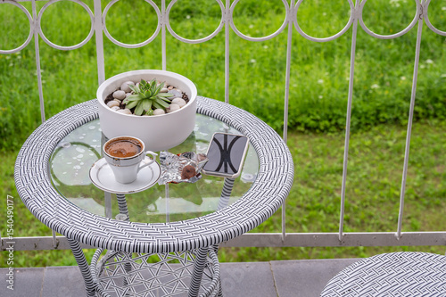 A cup of coffee, a smartphone and a potted houseplant on a glass table on a balcony.