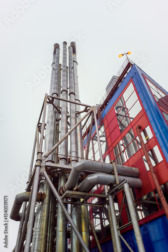 Tall building with a red and blue facade and a yellow roof. The building is surrounded by pipes and has a large chimney. Concept of industrialization and the importance of the building in the area