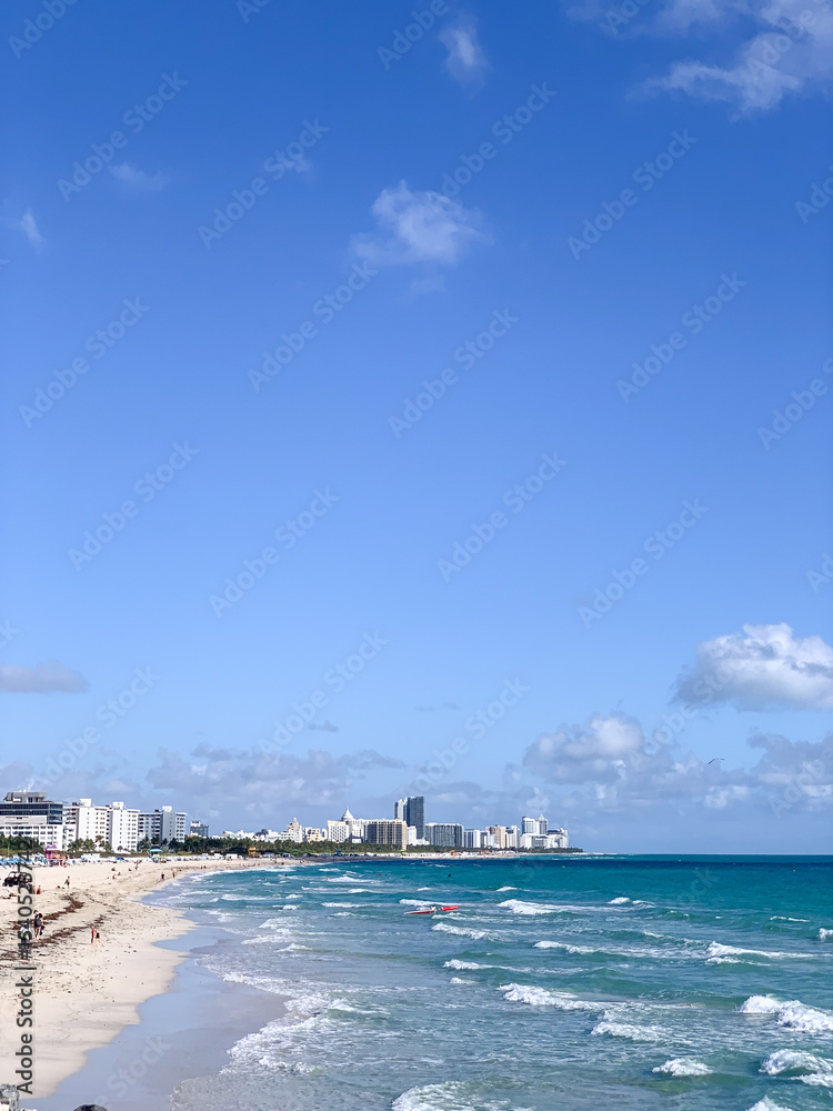 Fototapeta premium A bright, sunny view of Miami Beach with turquoise ocean waves, white sandy shore, and a backdrop of hotels and high-rise buildings along the coastline. 