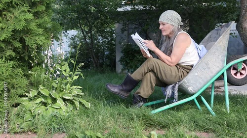  elderly woman  with long grey hair  sits in a garden wheelbarrow and and reads a book in the summer garden near the house. 