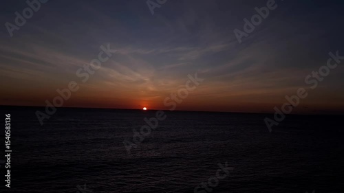 The glowing orange sun touches the ocean’s edge in slow motion, under a softly clouded sky, as the sea below darkens and settles into calm.
