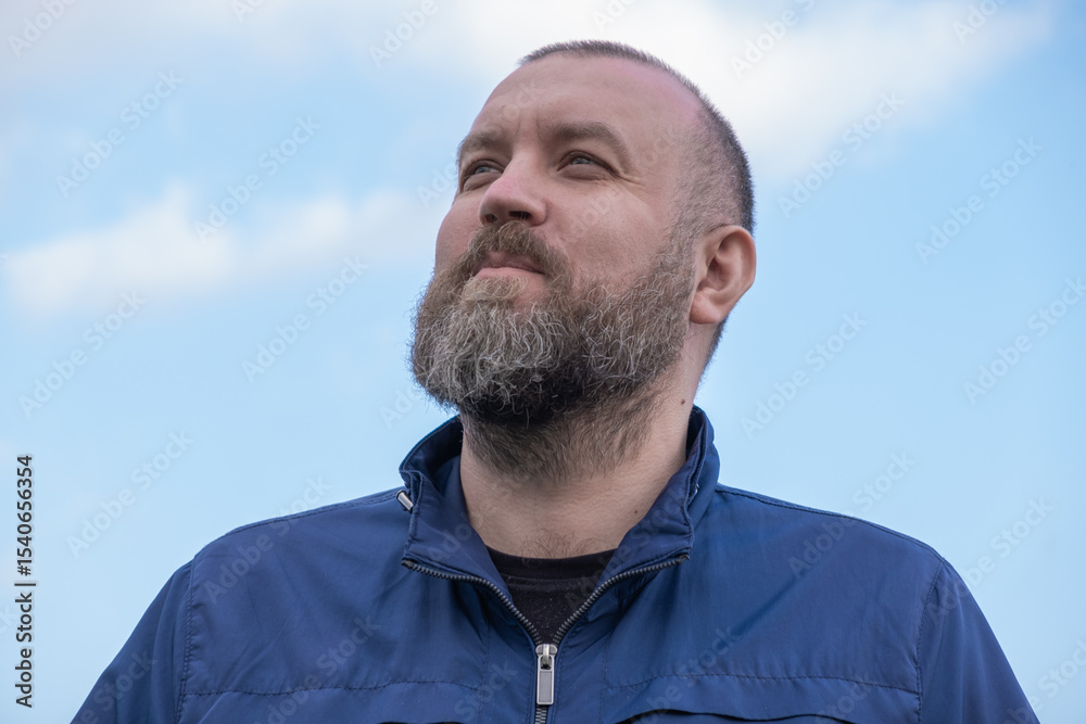 Fototapeta premium Man with beard gazes upward against a blue sky in a serene outdoor setting in daytime