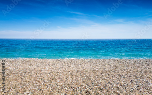 Pebble beach stretches along calm turquoise sea under bright blue sky. Sea and beach meet at clear horizon on sunny summer day. Peaceful coastal landscape without people or buildings