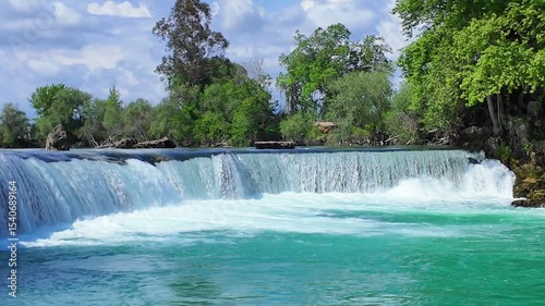 Calm flowing Manavgat waterfall with sparkling water descending over layered stones surrounded by lush forest greenery under warm sunlight on clear summer day, close up