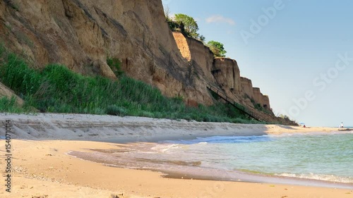 A wide sandy beach at the base of dramatic, high clay cliffs, with gentle waves breaking on the shore and a clear blue sky above.
