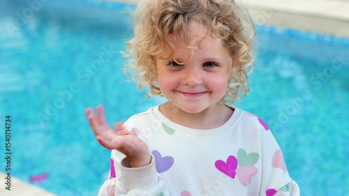 A smiling child with blonde curly hair, wearing a heart-patterned top, waves at the camera while standing in front of a blue swimming pool.
