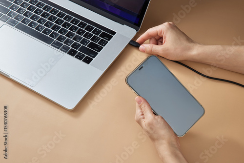 female hand connecting charger cable to laptop and holding smartphone at beige table, closeup