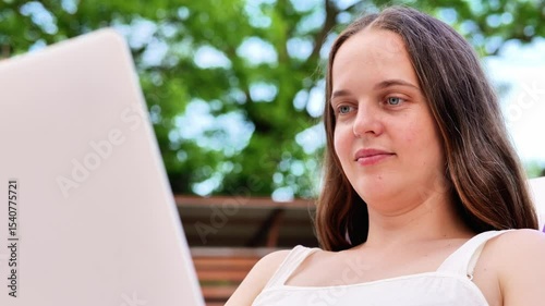 A young woman with long dark hair and blue eyes looks thoughtfully at a laptop screen, with a blurred green background, possibly outdoors.