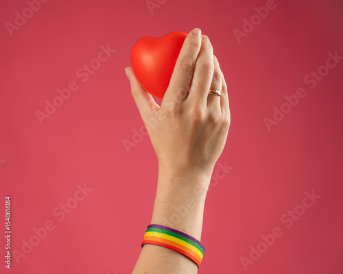Woman with engagement ring and rainbow bracelet holding heart on pink background. 