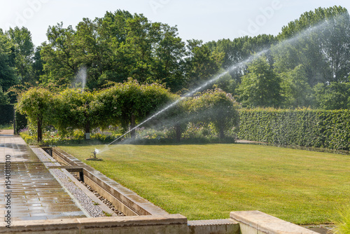 An automatic lawn sprinkler waters a well-kept garden in the sunshine. Hedges and pergolas with climbing plants grow in the background.