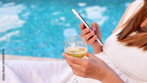 A person holds a white smartphone in one hand and a glass of white wine in the other, with a bright blue swimming pool as a blurred background.
