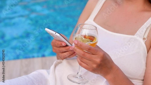 A woman in white poolside holds a smartphone and a glass of refreshing beverage, embodying summer relaxation and connectivity.