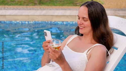 A happy young woman relaxes by the pool on a lounge chair, holding a smartphone and a drink, enjoying a perfect summer day.