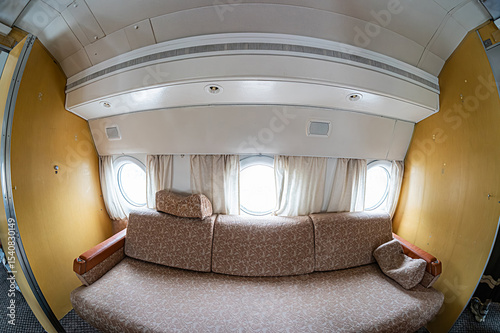 Luxurious airplane cabin with curved design, cream walls and ceiling, beige-framed oval windows, floral browntan sofa, wooden armchair, round side table, warm diffused lighting