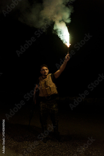 Soldier wearing a vest and military equipment, at night, alone, holding a machine gun in one hand and a flare in the other, which illuminates the area a little so his unit can locate him.