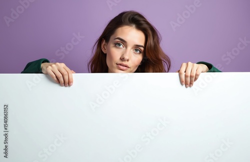 Attractive young woman peeks behind white empty banner on violet background. Stylish casual green clothes. Advertisement promotion copy space. Female student with happy smile makes announcement. photo