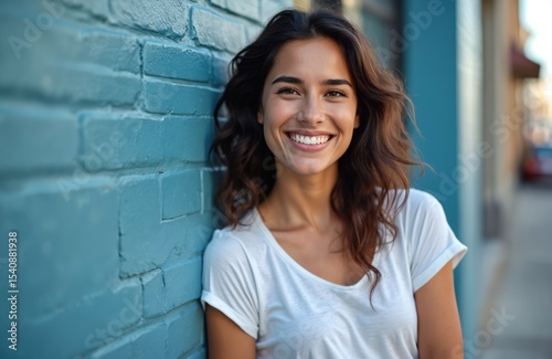 Beautiful hispanic woman smiles outdoors. Happy brunette girl smiles confidently, leaning turquoise wall. Attractive female with white t-shirt, sunlight on sunny day.