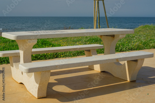Stone picnic bench and waste bin on a seaside promenade with clear blue sky and calm ocean in the background.