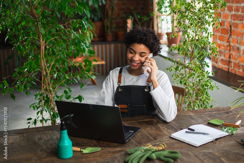 African woman florist gardener working in botanical store using laptop order plants by phone communicating with customer. Female small business owner working at flower shop. Running of own business