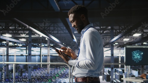 Photography Technician walks in data center, doing maintenance tasks on tablet, ensuring optimal performance
