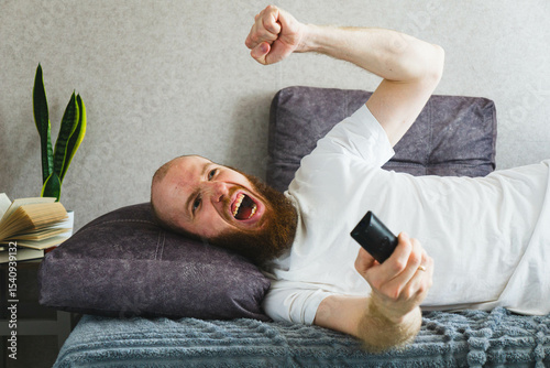 Happy bearded man lying on the bed with a remote control, laughing and cheering. Concept of joy, relaxation, and fun leisure time at home.

