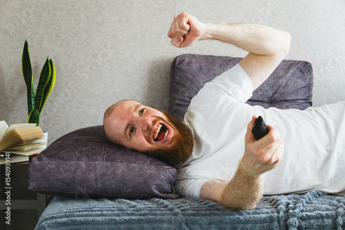 Happy bearded man lying on the bed with a remote control, laughing and cheering. Concept of joy, relaxation, and fun leisure time at home.

