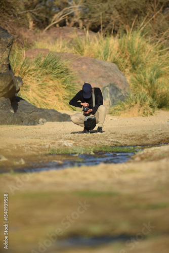 Unrecognizable man crouching by a stream, taking photographs with a DSLR camera, wearing a black cap and shoulder bag, surrounded by rocks, bushes, and sunlight in a natural outdoor setting.