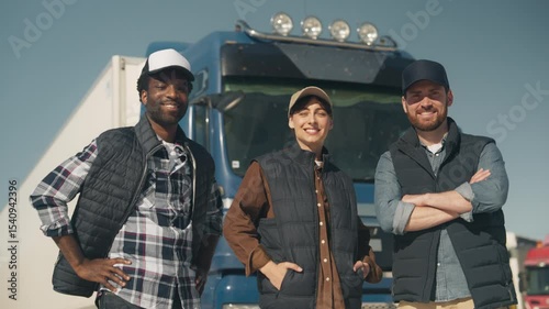 Three proud logistics workers looking directly at camera, standing confidently in front of parked blue truck. Wearing casual outdoor vests and caps, posing as a team under clear daylight.