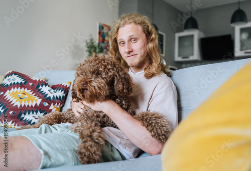Portrait of young teenager boy with long curly hear smiling cheerfully sitting on comfortable living room sofa hugging Maltipoo dog showing joy of bonding with beloved pet