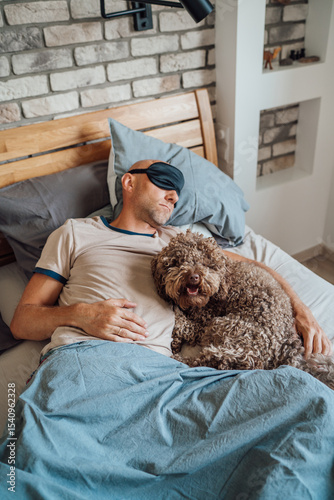 Maltipoo dog in bed with owner wearing black sleep eye mask during daytime nap on unfocused background. Funny pets and healthy lifestyle concept image.