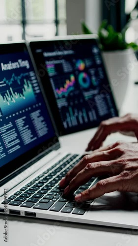 Close-up, side angle of hands typing on a laptop with financial graphs on screens, conveying a modern tech workspace, ideal for a business video.