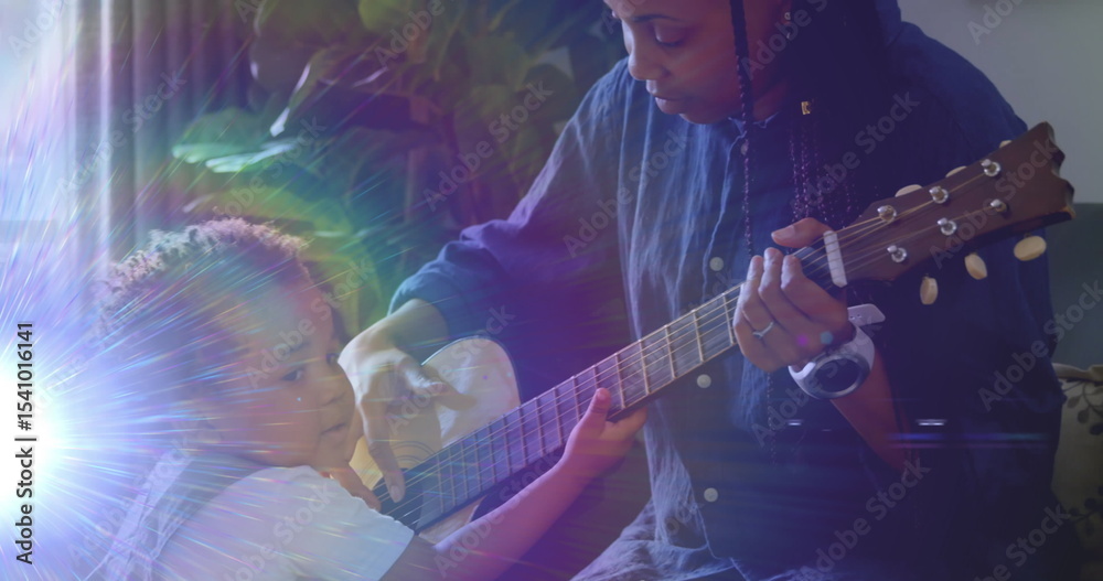 Obraz premium Mother in blue shirt guiding toddler strumming acoustic guitar near living room window, with plant