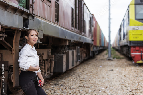 A female professional stands beside a freight train holding a white safety helmet and keys. She appears to be observing or supervising logistics activities in a rail transport environment.