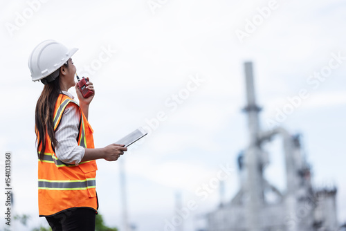 A female engineer in a safety vest and helmet communicates via walkie-talkie while holding a tablet. She observes operations at an industrial oil refinery site in the background.
