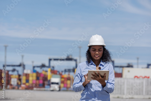 A female manager wearing a helmet and holding a tablet points forward at a busy shipping port. Surrounded by containers and trucks, she oversees logistics and directs port operations.