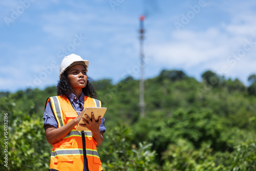 A female environmental engineer in safety gear takes notes outdoors near a forested area with a visible industrial chimney, indicating monitoring or assessment of air pollution impact.