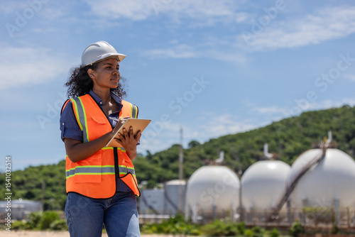 A female engineer in safety gear surveys a site with large spherical chemical tanks. Holding a tablet, she conducts an inspection or environmental assessment in an industrial storage area.