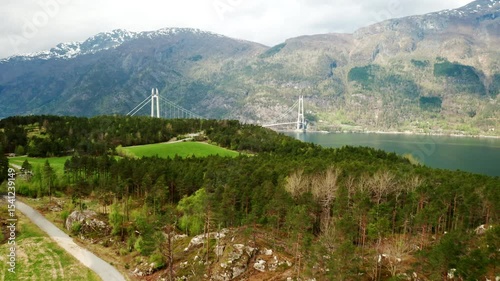 Majestic Hardanger Bridge spans wide body of water nestled among lush hills and mountains. Suspension bridge elegant towers stand out in landscape