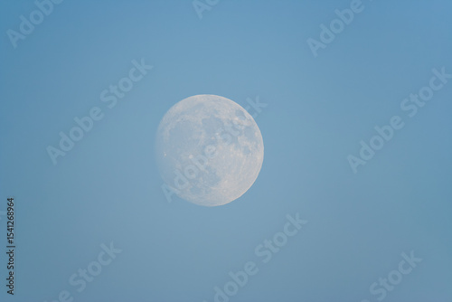 The full moon visible in broad daylight against a clear blue sky.