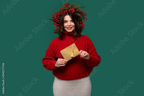 Cheerful woman in a red sweater holding a golden envelope, wearing a berry wreath, smiling