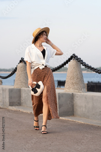 Woman walks stylishly holding handbag at sunset along promenade.